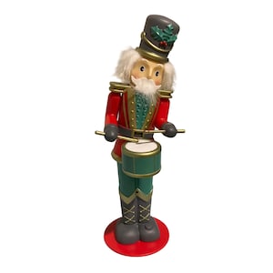 May include: A nutcracker figurine wearing a grey hat with holly leaves, a red jacket with gold trim, and green trousers. The figure has white hair and a beard, and is holding two drumsticks over a green drum with gold accents.