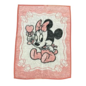 May include: A rectangular pink and white towel featuring a graphic of a baby Minnie Mouse character holding a heart-shaped rattle. The towel has a decorative border with white bows in each corner and a solid pink trim around the entire edge.