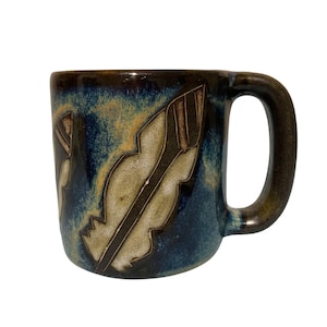May include: A ceramic mug featuring a deep blue and brown glaze with a mottled texture. The exterior surface is decorated with two carved, light-colored feather motifs that contrast against the dark, blended background colors of the pottery.