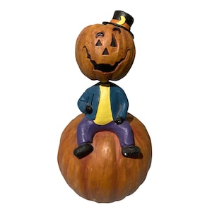 May include: A decorative figurine featuring a carved pumpkin head with a smiling face wearing a small black top hat with a yellow crescent moon. The figure has a yellow shirt, blue jacket, and purple pants, and is sitting on a large orange pumpkin.
