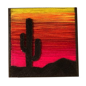 May include: A square string art piece featuring a black silhouette of a saguaro cactus and desert hills against a vibrant gradient background of horizontal threads transitioning from yellow at the top to orange, red, and bright pink at the bottom.