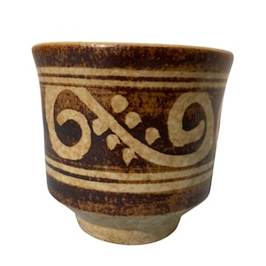 Op de afbeelding: Een kleine keramische beker met een bruine buitenkant en een beige decoratief patroon. De beker heeft een afgeronde body en een voet. Het ontwerp omvat gebogen lijnen en stippen. De beker wordt waarschijnlijk gebruikt om te drinken.
