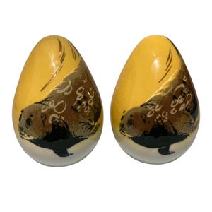 May include: Two egg-shaped ornaments featuring a yellow background with a painted brown and black sea otter illustration. The design includes stylized white circular patterns and gold-toned accents on the upper portion of each smooth, rounded object.