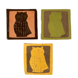 May include: Three square ceramic tiles featuring owl silhouettes. One tile has a black owl on a terracotta background, one has a lime green owl on a dark gray background, and one has a yellow owl on a dark gray background. Each owl has a grid pattern.