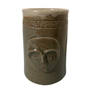 Peut inclure: Un vase cylindrique en céramique dans les tons gris et marron. Le vase présente un motif de visage sculpté et le mot "WALK-ELA" autour du haut. Le vase a une surface texturée et un bord de couleur claire.