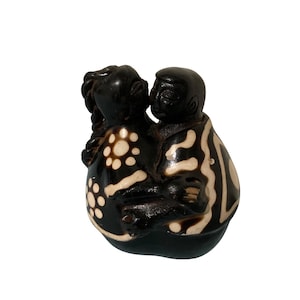 Puede incluir: Una escultura marrón oscuro que representa a dos figuras abrazándose. Una figura tiene un peinado trenzado y un patrón de círculos blancos. La otra figura tiene un diseño estampado de líneas blancas. La escultura está sobre un fondo blanco.