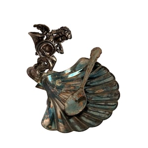 May include: Silver-toned seashell-shaped dish with a small spoon and a cherub figure playing a horn. The dish has a ribbed texture and a tarnished appearance, suggesting an antique or vintage item.