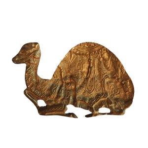 May include: A detailed, golden-colored metal camel figurine. The camel is depicted in a seated position with intricate embossed patterns on its body and saddle. The figurine has a detailed head and neck, and is set against a white background.