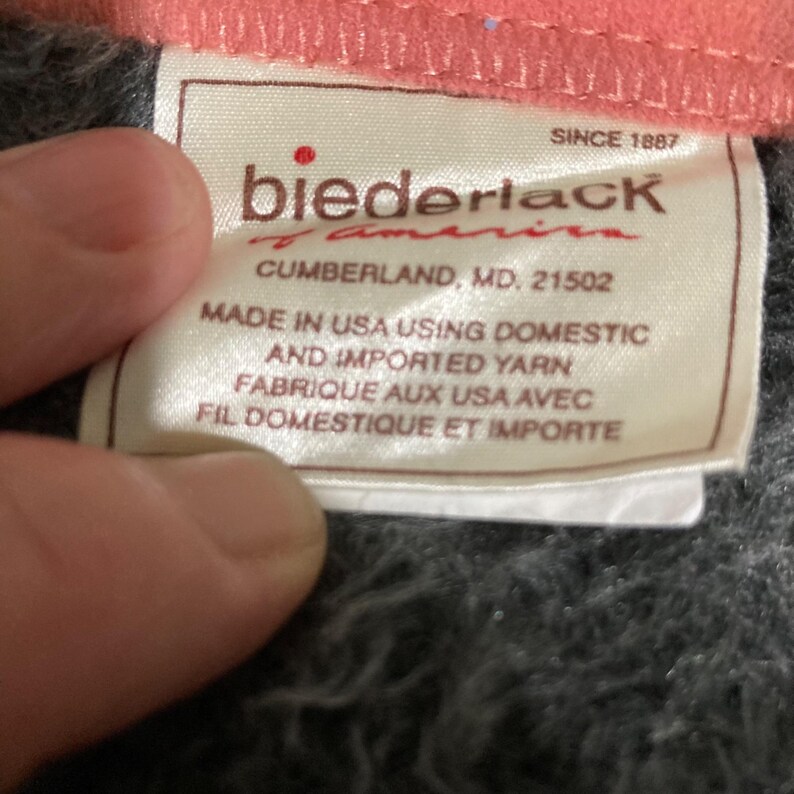 May include: A white fabric tag with brown text attached to a dark gray textured blanket. The tag reads: Biederlack of America, Cumberland, MD. 21502, Made in USA using domestic and imported yarn, Fabrique aux USA avec fil domestique et importe.