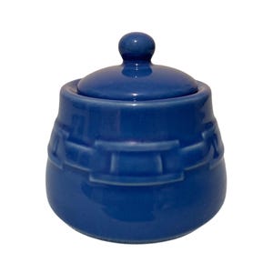 Puede incluir: Un azucarero de cerámica azul sólido con una tapa a juego que presenta un pequeño pomo redondo como asa. El exterior del cuenco está decorado con un patrón de banda horizontal en relieve que rodea el centro del recipiente redondeado.