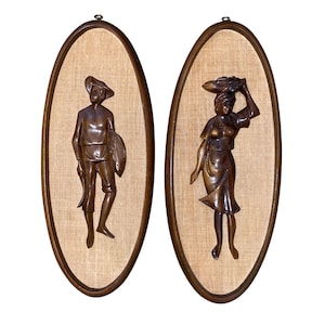 Op de afbeelding: Een paar ovale wandplaten met donkerbruine uitgesneden houten figuren tegen een lichtbeige achtergrond van gestructureerde stof. De ene plaat toont een figuur met een hoed en een bundel, de andere een figuur die een mand draagt.