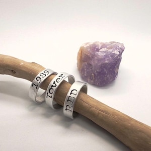Könnte beinhalten: Drei silberne Ringe mit den Worten "1989", "Lover" und "RED" auf einem Holzstab. Im Hintergrund befindet sich ein violetter Amethystkristall. Die Ringe sind wahrscheinlich Schmuck.