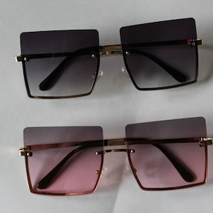 May include: Two pairs of square-shaped sunglasses with a gold frame and black arms. The top pair has a gradient lens that transitions from dark gray to light gray. The bottom pair has a gradient lens that transitions from dark pink to light pink.