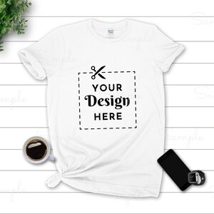 May include: White t-shirt with the text "YOUR Design HERE" inside a dashed square. The shirt is styled with a knot at the bottom. A cup of coffee, a smartphone, and a smartwatch are placed near the shirt.