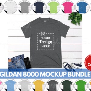 May include: A mockup bundle of 41 Gildan 8000 t-shirts in various colors. The t-shirts are laid out on a white wooden surface with a pair of jeans, a phone, and a plant. The t-shirt in the center has the text "Your Design Here" on it.