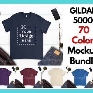 May include: A mockup bundle of 70 different colored Gildan 5000 t-shirts. The t-shirts are laid out on a white wooden surface with jeans, a phone, and a plant. The text "YOUR DESIGN HERE" is on the front of the navy blue t-shirt.