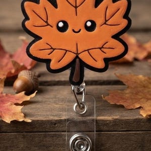 May include: A badge reel featuring a bright orange maple leaf with a black outline and a cute, smiling face with two black eyes. The leaf is attached to a clear plastic strap with a metal snap button and a retractable cord mechanism.