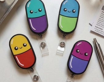 May include: Four oval-shaped badge reels featuring cartoon pill designs with smiling faces. Each pill has a two-tone color scheme: teal and purple, blue and green, yellow and red, and purple and light purple. Each reel has a clear plastic strap.