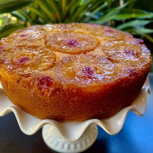 Può includere: Una torta rovesciata all'ananas di colore marrone dorato con una glassa lucida, appoggiata su un supporto per torte bianco.