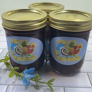 Op de afbeelding: Drie glazen potten gevuld met een donkerpaarse jam. Elke pot heeft een gouden deksel en een blauw en wit etiket met de tekst "A Slice of The Virgin Islands".