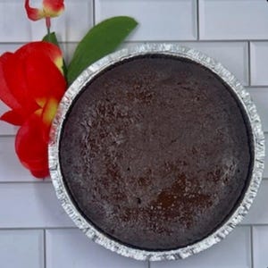 May include: A round, dark brown cake in a silver foil pan. A vibrant red flower with green leaves is placed to the left of the cake. The cake sits on a white tiled surface.