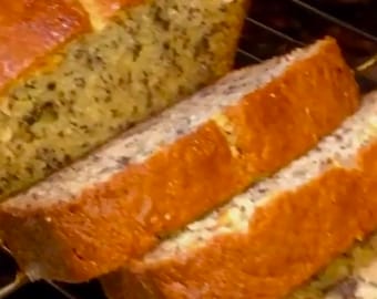 Può includere: Una pagnotta di banana bread tagliata a fette su una griglia. Il pane è dorato e ha un aspetto maculato dovuto alla banana e alle noci.