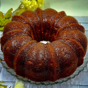 May include: A brown bundt cake with a glazed finish, sitting on a clear glass plate. The cake is on a white tiled surface with a few yellow flowers in the foreground.
