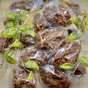 May include: A pile of individually packaged brown crumbly treats, likely a type of granola or cookie, wrapped in clear plastic bags with gold ribbon ties. The treats are arranged on a light brown surface with green plastic leaves scattered around.