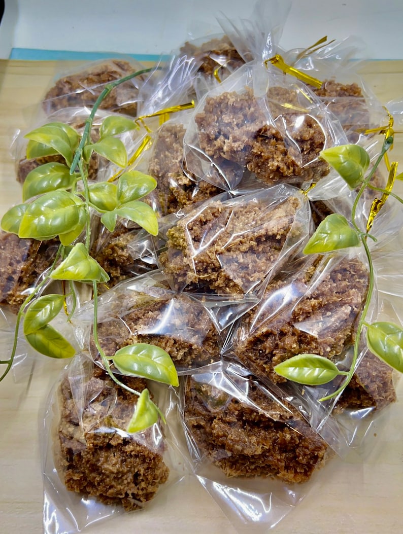 May include: A pile of individually packaged brown crumbly treats, likely a type of granola or cookie, wrapped in clear plastic bags with gold ribbon ties. The treats are arranged on a light brown surface with green plastic leaves scattered around.