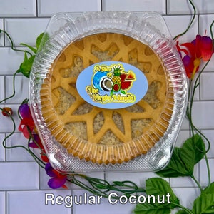 May include: A round coconut pie with a lattice crust in a clear plastic container. The pie is topped with a light brown coconut filling. The container has a sticker that says "A Slice of The Virgin Islands".