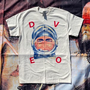 May include: A white short-sleeve t-shirt featuring a graphic of a person wearing a blue astronaut helmet. The red letters D, V, E, and O are positioned around the helmet graphic, with D and V above and E and O below the central illustration.
