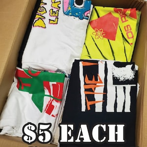 May include: A cardboard box filled with five folded t-shirts. The shirts have various colorful graphic designs and text, including "Digital Leaf", "Bad So", "The", and an atom symbol. The box has a sign that reads "Misprint Sale $5 Each + Shipping".