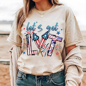 以下が含まれることがあります： ベージュ色のTシャツ。青い筆記体で「let's get LVT」と書かれ、愛国的なグラフィックが描かれています。デザインには、花火、星、アメリカ国旗模様のロケットが含まれており、独立記念日の祝賀に最適です。