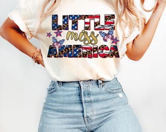 May include: Cream-colored t-shirt with the text "Little Miss America" in a patriotic design. The text features stars, stripes, and leopard print accents, along with butterfly and star graphics. The person is wearing light blue jeans.