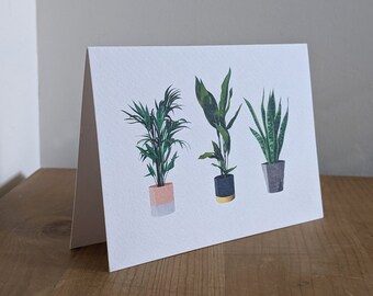 May include: A greeting card with a white background featuring three potted plants. The plants are a green palm, a green plant with a dark gray pot, and a green snake plant with a light gray pot.