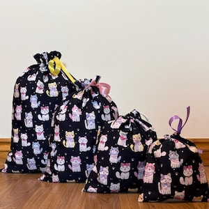 May include: Four drawstring bags in different sizes, featuring a repeating pattern of cartoon cats in various colours on a black background. The bags have yellow, pink, and purple ribbons.