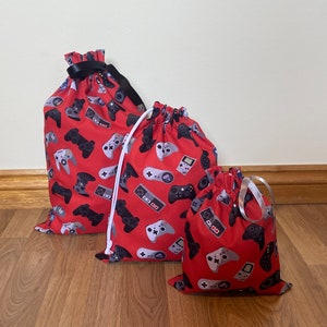May include: Three red drawstring bags featuring a pattern of grey and black video game controllers. The largest bag has a black ribbon closure, the others have white and silver cord closures. The bags are different sizes.