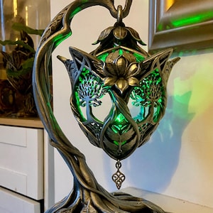 May include: A decorative table lamp with a nature-inspired design. The lamp features a bronze-coloured metal frame shaped like a tree branch, supporting a floral-shaped shade. The shade has cut-out tree designs and emits a vibrant green light.