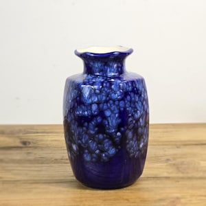 May include: A ceramic vase with a deep blue base color and a mottled, crystalline glaze pattern in lighter shades of blue and white. The vase features a rounded body, a narrow neck, and a flared, wavy rim, showcasing a glossy, smooth finish.