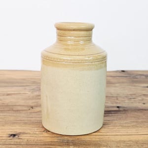 Op de afbeelding: Een cilindrische stenen kruik met een tweekleurig beige en lichtbruin glazuur. De pot heeft een smalle hals met horizontale ribbels en een decoratieve reliëfband rond de schouder, die overgaat in een glad, lichter gekleurd lichaam.
