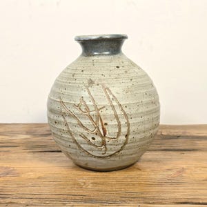 May include: A round ceramic vase with a speckled off-white glaze and horizontal ribbed texture. The vase features a dark blue-grey rim and a hand-etched abstract leaf design on the front. The body is spherical with a narrow neck and opening.