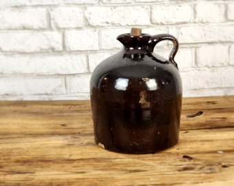 May include: A dark brown glazed ceramic jug with a rounded body, a short neck, and a single side handle. The jug features a small spout and a light-colored cork stopper inserted into the opening, with visible surface wear and minor chips.