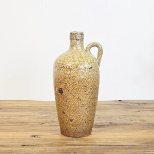 May include: A tall, cylindrical stoneware jug with a narrow neck and a single side handle. The exterior features a mottled, speckled brown and tan glaze with a lighter, cream-coloured textured finish around the upper shoulder and neck area.