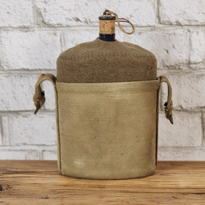 May include: A brown canvas canteen with a brown felt cover and a cork stopper. The canteen has a brown leather strap and a brass buckle.