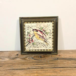 May include: A square decorative tile featuring a yellow and brown bird perched on a branch with pink flowers, set within a dark metallic frame. The tile has a cream-coloured background with a black and white checkered border around the illustration.