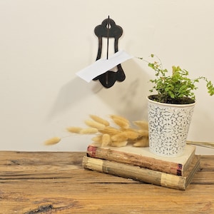 May include: A black metal wall-mounted mail holder with a white paper. A small potted plant with green leaves sits on two old books. Dried wheat and a wooden table complete the scene.