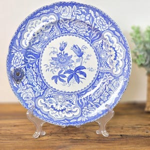 May include: A round ceramic plate featuring a blue and white floral pattern. The center displays a detailed botanical illustration of a passion flower, while the border is decorated with intricate rose motifs and ornate scrollwork designs.