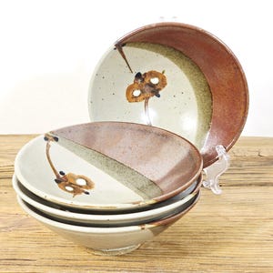 May include: A set of four ceramic bowls with a unique design. The bowls feature a cream-colored base with brown and green accents, and a floral motif. The bowls are stacked on a wooden surface.