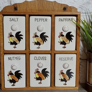 Può includere: Un porta spezie in legno con sei barattoli in ceramica bianca. Ogni barattolo ha un'illustrazione di un gallo e un'etichetta con il nome di una spezia: Sale, Pepe, Paprika, Noce moscata, Chiodi di garofano e Riserva.