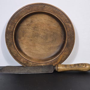 May include: A wooden bread plate and bread knife. The plate is round with a raised rim. The knife has a wooden handle with the word "BREAD" carved into it. Both are a warm brown colour.